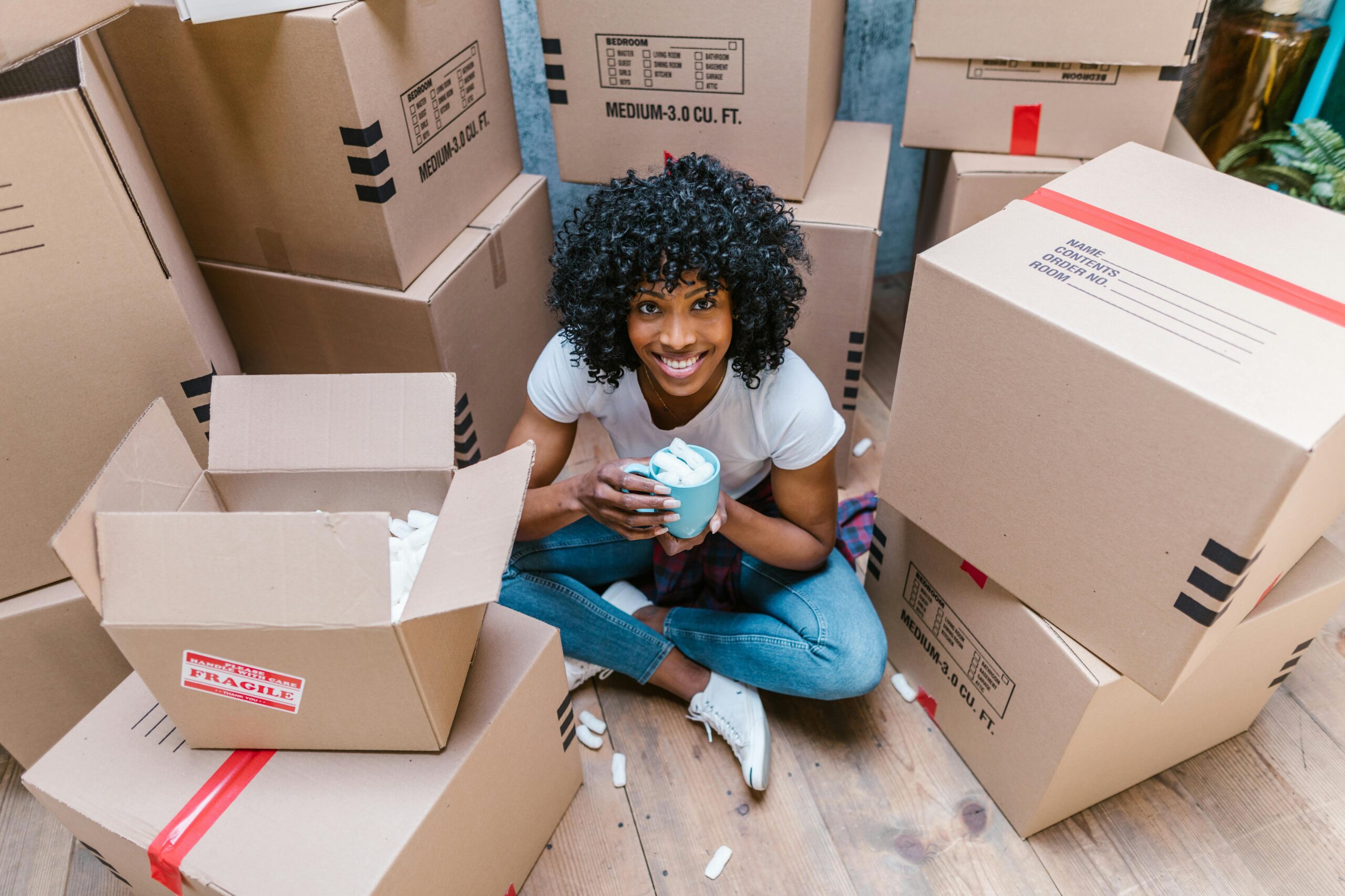 A woman sitting among moving boxes with a mug, surrounded by packing materials.