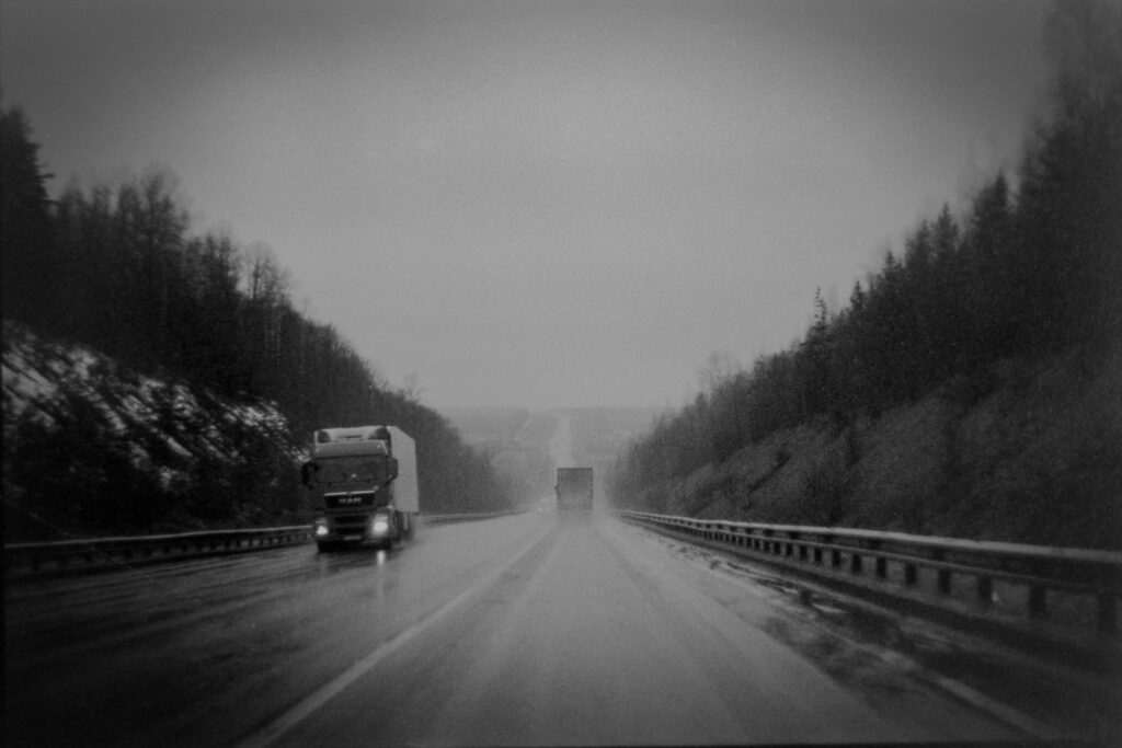 Black and white photograph of trucks traveling on a forest highway in gloomy weather.